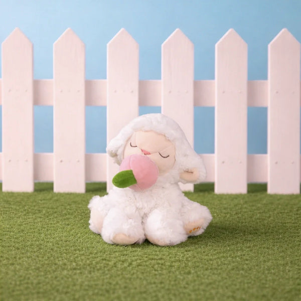Plush toy sheep with a pink bubble gum sitting on grass with a white picket fence in the background