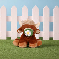 Plush toy cow with a flower on its face in front of a white picket fence.