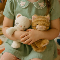 Child holding two plush cats, one white and one orange, wearing a green dress with cat-themed collar.