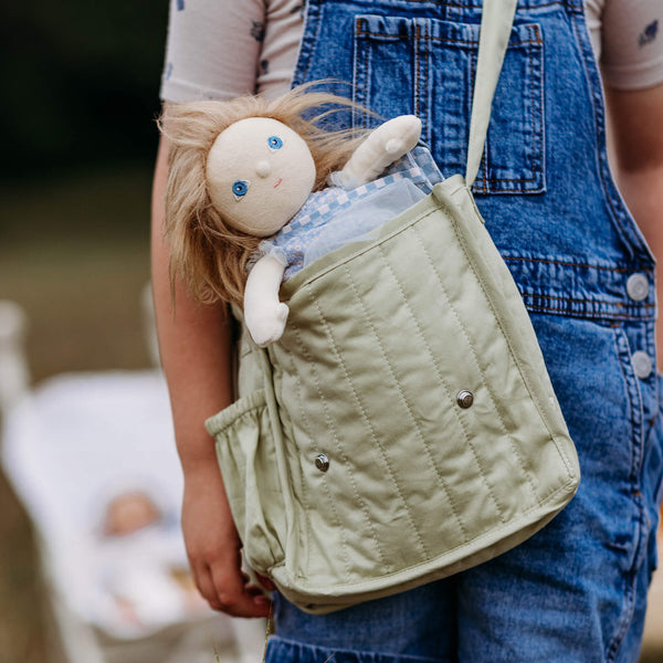 Person holding a green quilted bag with a doll inside, wearing blue overalls.