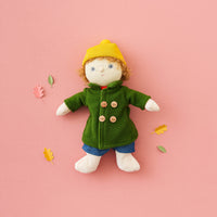 Toy figure wearing a green coat and yellow hat on a pink background with autumn leaves.