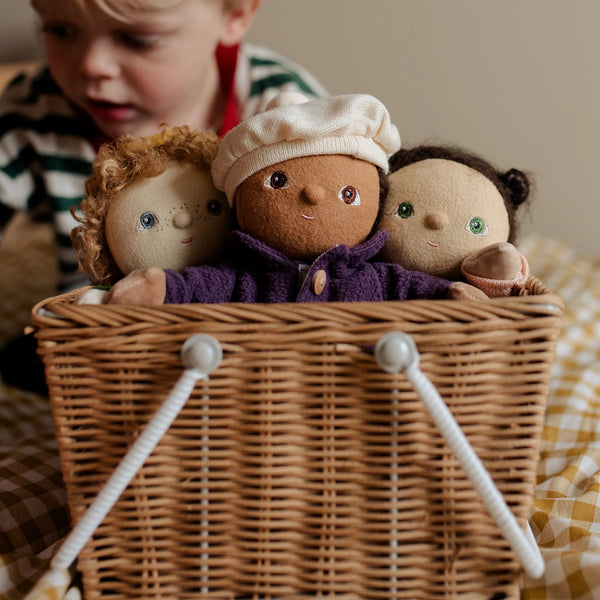 Three dolls with different hair colors and outfits inside a wicker basket, with a child in the background.
