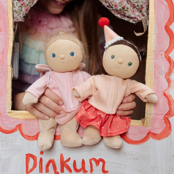 Two dolls held by a person with a decorative background and 'Dinkum' branding.